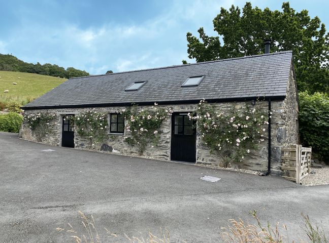 A stone building with two black doors two windows and climbing roses along the boundary at The Shippon in Llanrwst