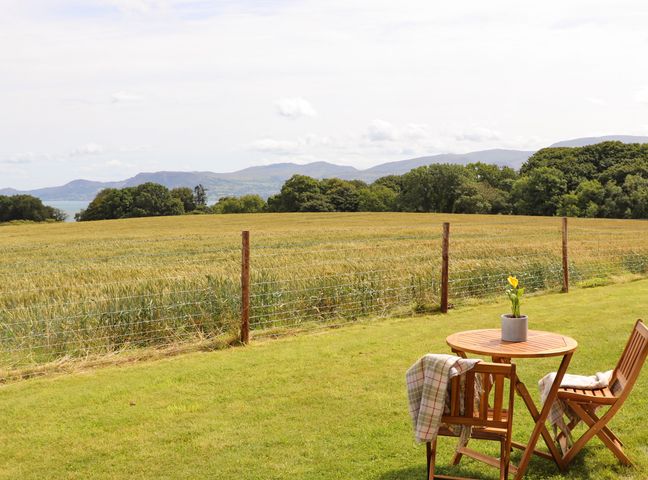 A wooden table and two chairs on grass overlooking a fenced field with hills and trees in the background at Bwthyn Cae Haidd in Beaumaris