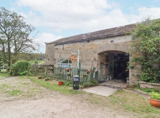 Exterior of a stone barn with an arched entrance and a green picket fence with flower pots at Moldy Warp Barn in Haworth