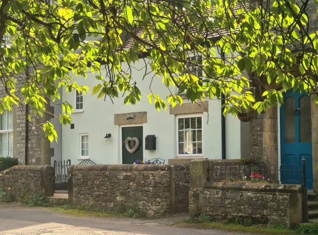 A stone wall and green tree branches in front of a white house with a dark green door and window at Church View Cottage in Litton Derbyshire