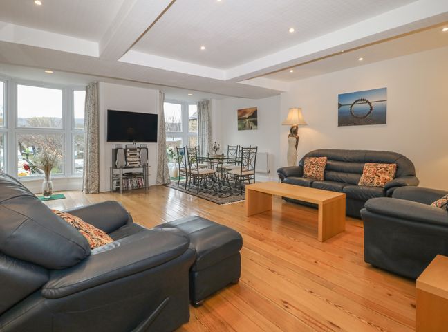 A living room with three blue sofas a wooden coffee table a glass dining table with six chairs a television and large windows at Park Road Apartment in Pateley Bridge