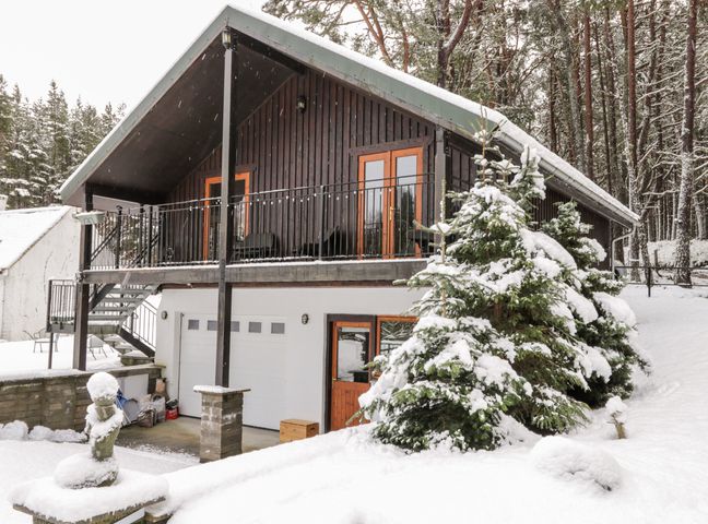 A snowy house with a balcony and garage doors surrounded by snow-covered trees at Torbreck Chalet in Kincraig