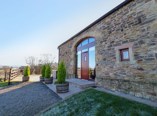 The stone exterior of a building with a wooden door and large window surrounded by wooden planters with small trees at Hill Brook Barn in Chatburn