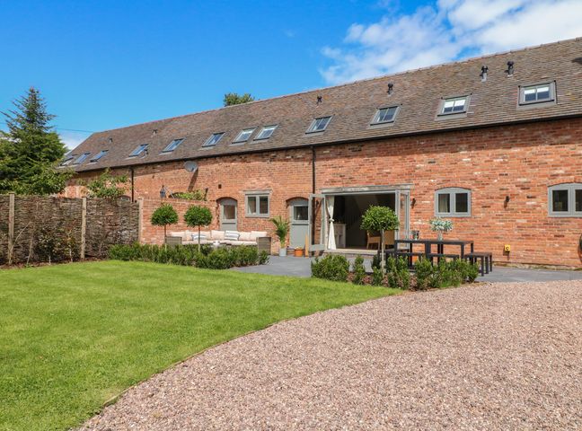 An outdoor patio area with seating and a dining table next to a long brick building with multiple small windows and skylights at Upper Barn in Great Haywood