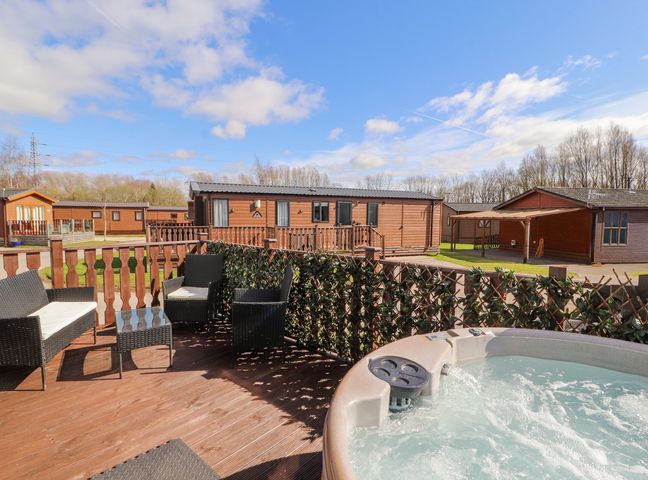 An outdoor deck with a hot tub and wicker chairs surrounded by wooden fences and lodges at Duckling Lodge South Lakeland Leisure Village