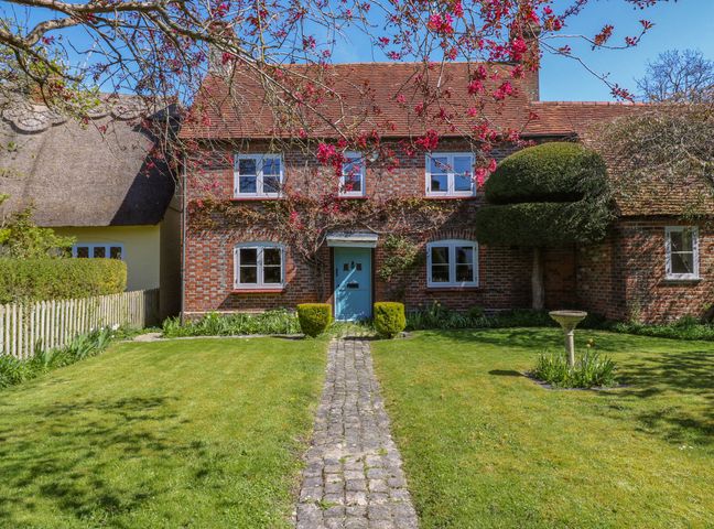 A brick cottage with blue door fronted by lawn and stone path with trimmed hedge and blossom tree branches at Bakehouse Farm in Bledlow