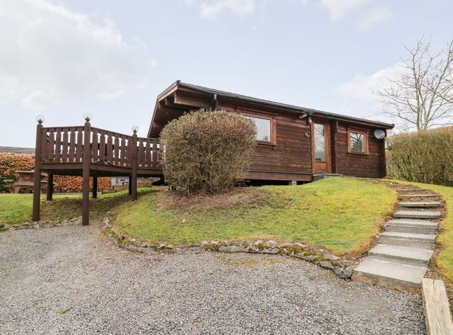 A wooden cabin with a deck on a grassy hill with stone steps and a gravel driveway at Snowy Owl Lodge in Rhayader