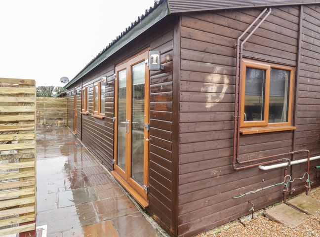 An exterior view of a brown wooden building with multiple windows and doors on a wet stone pavement at Sparrows Den Lodge in Winchelsea Beach