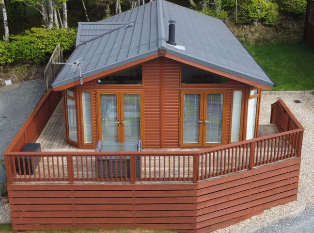A wooden lodge with a gray roof surrounded by a wooden deck and fence in a green area at Lodge 11 in Tavistock