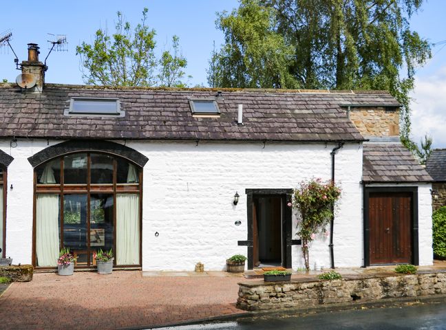 A white stone house with large windows and a wooden door surrounded by plants at Smithy Cottage in Wigglesworth near Long Preston