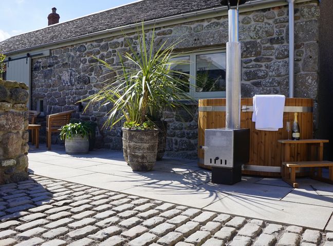 An outdoor patio with a cobblestone and paved floor a wooden hot tub with a towel and champagne bottle a potted plant and wooden chairs by a stone house at The Wink Cape Cornwall