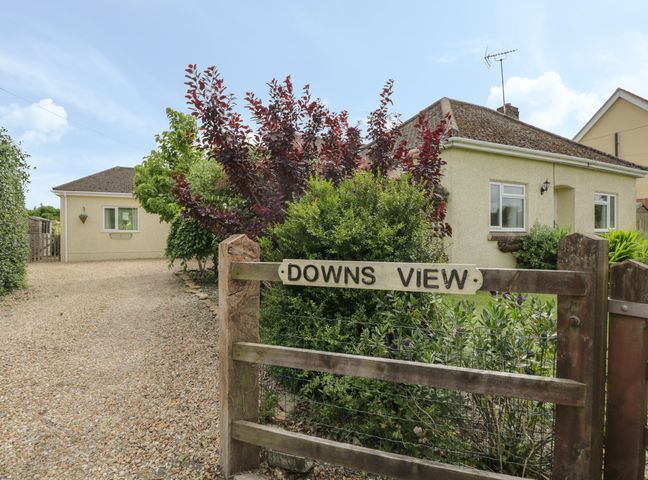 A gravel driveway leading to a single-story house with bushes and a wooden gate with a sign that says Downs View at Norton Ferris