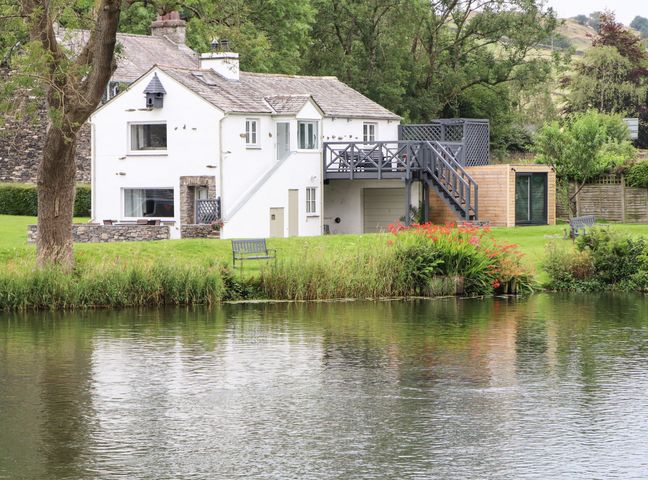 A white house with a deck and stairs by a pond with bench and plants at Riverside Retreat in Newby Bridge