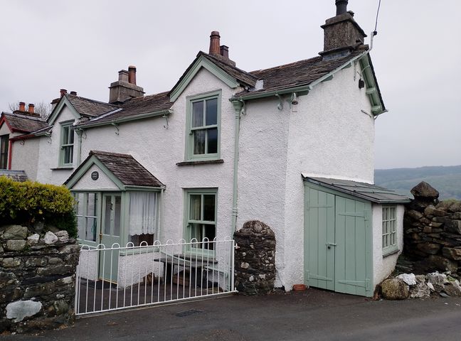 A white cottage with green windows and doors next to a stone wall at Sunbeam Cottage in Coniston