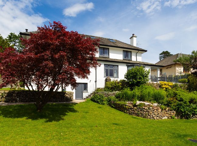 A white two-story house with black windows and a slate roof surrounded by a green lawn a red-leafed tree and stone garden walls at Wynford in Ambleside
