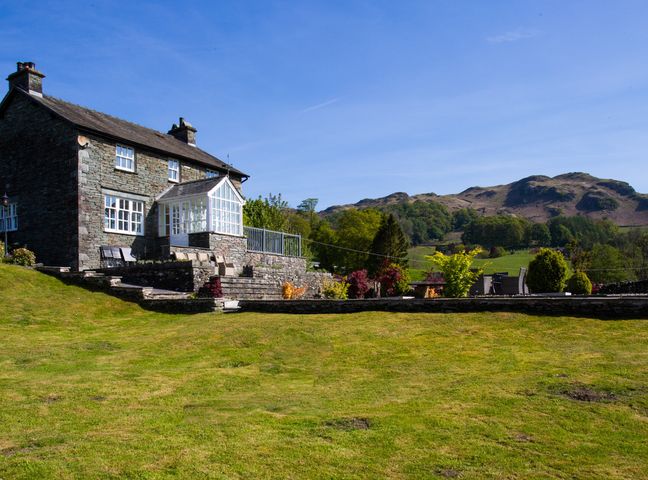 A stone house with a conservatory and garden furniture on a terrace with a lawn and hills in the background at Grass Ings in Langdale