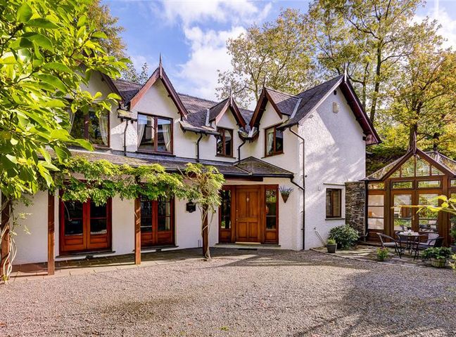 The exterior of a white house with wooden doors and windows with a gravel driveway and a glass conservatory at Silver Howe in Bowness