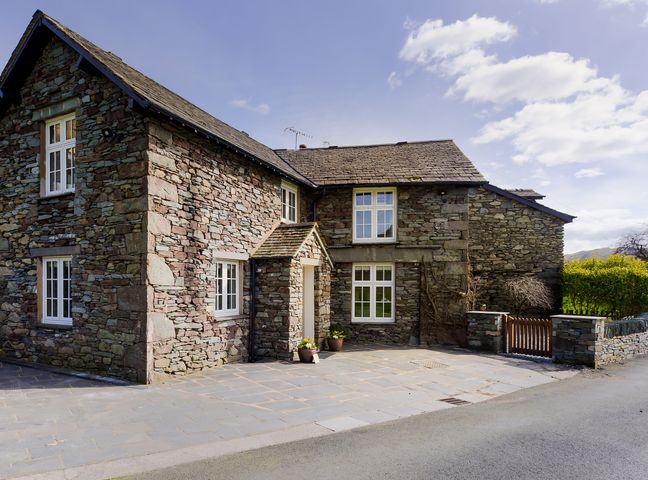 A stone house with white framed windows and a small porch on a paved driveway at Tilly's Cottage in Grasmere
