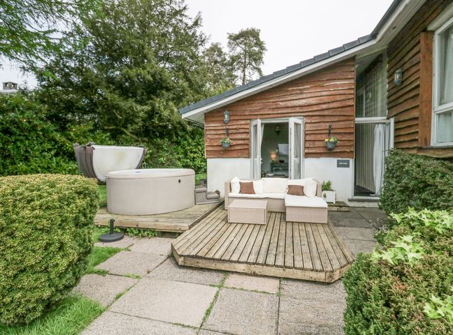 An outdoor patio with a wooden deck sofa hot tub and surrounding bushes at Lake Lodge in Bowness-On-Windermere