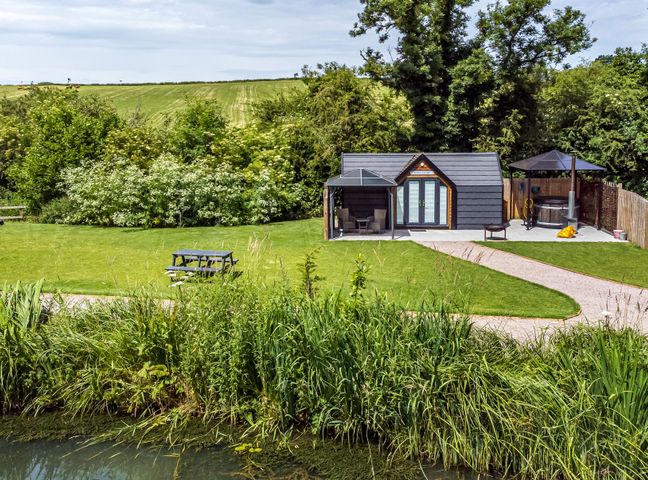 A small cabin with patio furniture and a hot tub beside a pond with grass and trees at Acorn Lodge At Bridge Lake Farm Fishery in Chacombe