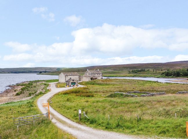 A gravel road leading to stone buildings near a body of water with grassy fields and hills in the background at Snave Barn Grimwith near Appletreewick