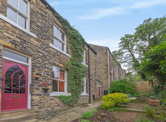 A stone house with a red door and ivy on the wall with a garden path and shrubs at Rose Cottage in Haworth