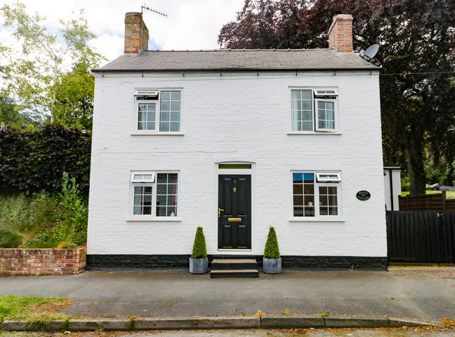 A white two-story house with four windows and a black door with two small plants in pots on the steps at Beech Cottage in Hunmanby