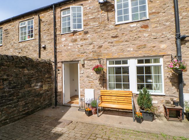 A stone exterior courtyard with a wooden bench plants and an open door at 1 The Stables in Alston
