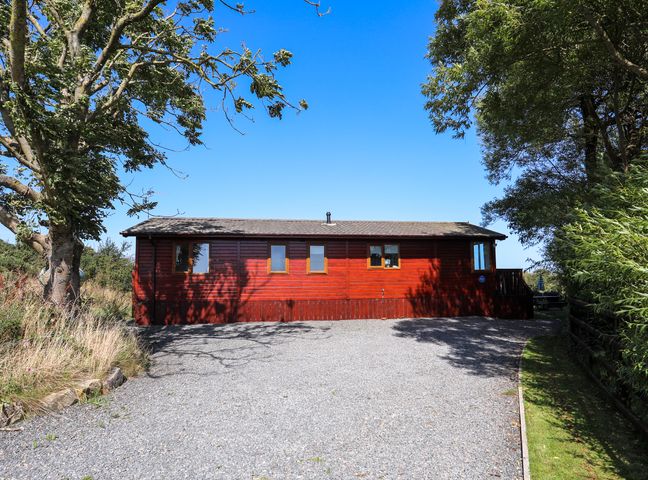 A red wooden cabin with multiple windows surrounded by trees and a gravel driveway at Ffrwd Lodge in Rhosneigr