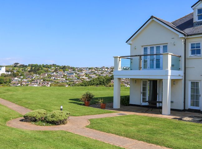 A two-story house with a balcony and patio on a lawn with a pathway and distant houses in South Milton near Thurlestone