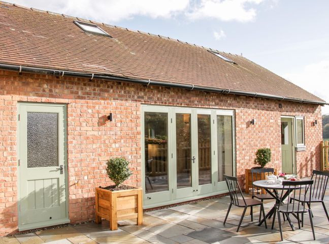 A brick building with green doors and windows a patio with a table chairs and potted plants at Conkers Cottage in Rushbury near Church Stretton