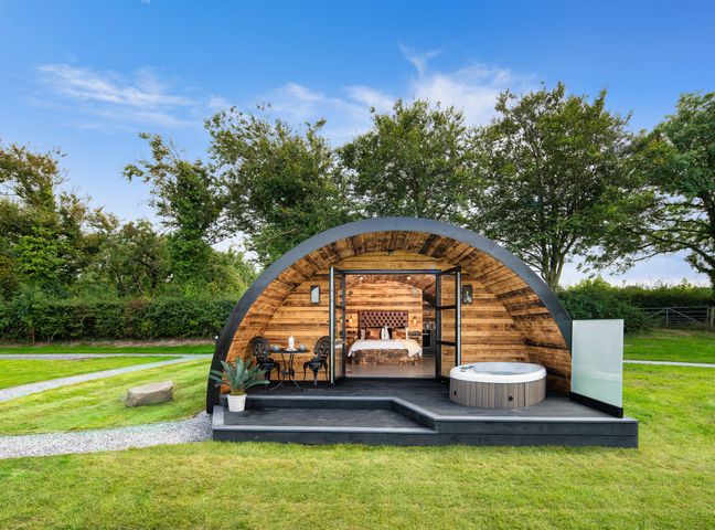 A pod-shaped wooden cabin with glass doors opening to a bedroom and outdoor seating with a hot tub at The Piglet Pod in Llanarth