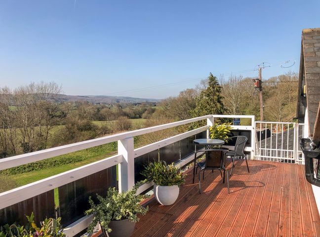 A balcony with potted plants a round table and two chairs overlooking trees and fields at Heather in Graigfechan