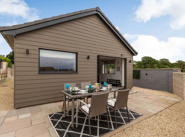An outdoor patio area with a dining table and six chairs beside a house with an open folding door at Secret Kingdom Cottage in Warkworth