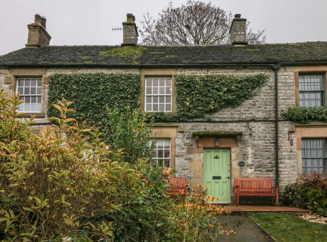 A stone cottage with a green door two wooden benches and plants in front at 3 Old Hall Cottages Monyash near Bakewell
