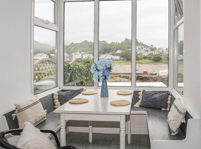 A dining nook with a white table and wicker placemats surrounded by black bench seating and cushions with a vase of blue flowers at Ivy Cottage in Borth-y-Gest near Porthmadog