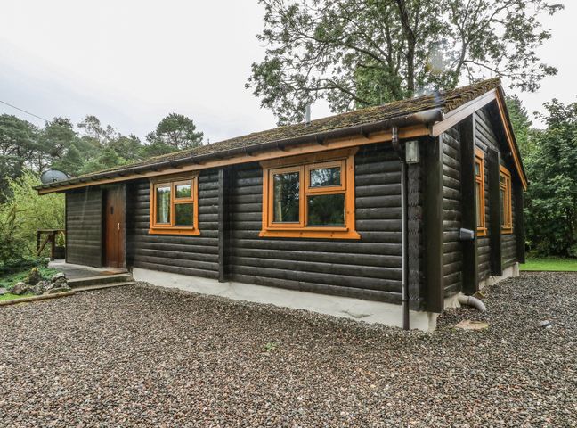 An exterior view of a dark wooden cabin with orange window frames and a gravel driveway at Millmore Cabin in Killin