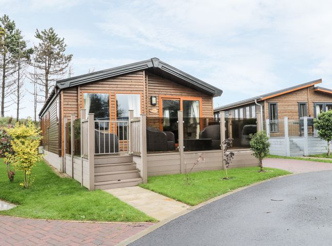 A wooden cabin with a raised deck and outdoor seating surrounded by grass and a paved road at Huckleberry in Runswick Bay near Staithes