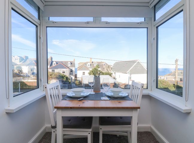A dining table set for two by a bay window with views of houses and the sea at The Beach Hut in St Ives