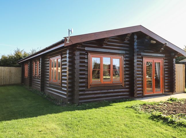 A log cabin with multiple windows and a glass door surrounded by grass and fenced yard at Rowan Lodge in Greetham Rutland