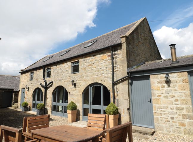 An outdoor patio area with wooden table and chairs in front of a stone building with arched windows and blue doors at Granary Cottage in Bellingham