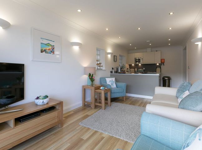 A living room with a sofa and armchairs a television on a wooden stand and an open kitchen at Atlantis in St Ives