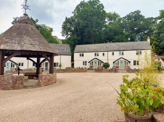 A courtyard with a thatched gazebo and two white cottages with green doors surrounded by trees at Pumphouse Cottage in Uffculme