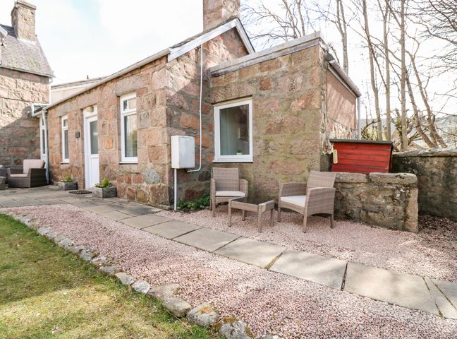 An outdoor stone patio area with two cushioned chairs and a small table beside a stone building at Gairnlea Cottage in Ballater