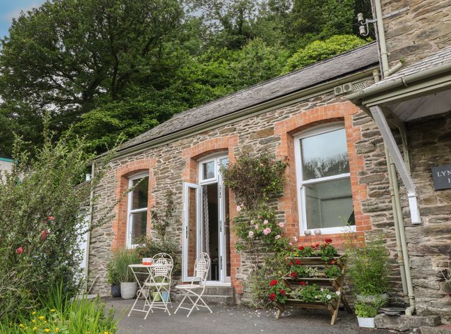 An outdoor patio area with two white metal chairs and a table in front of a stone building with red brick window frames and climbing plants at Ferndale in Lynton