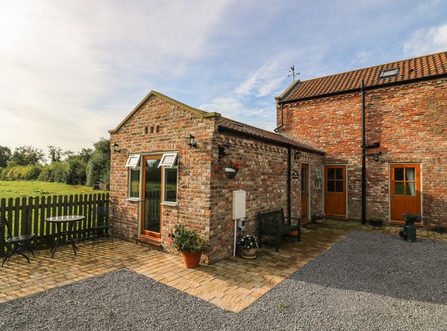 An outdoor area with brick buildings a wooden bench potted plants a round table with chairs and a fenced grassy field at Decca Cottage Nidd Grange Moor Monkton near York