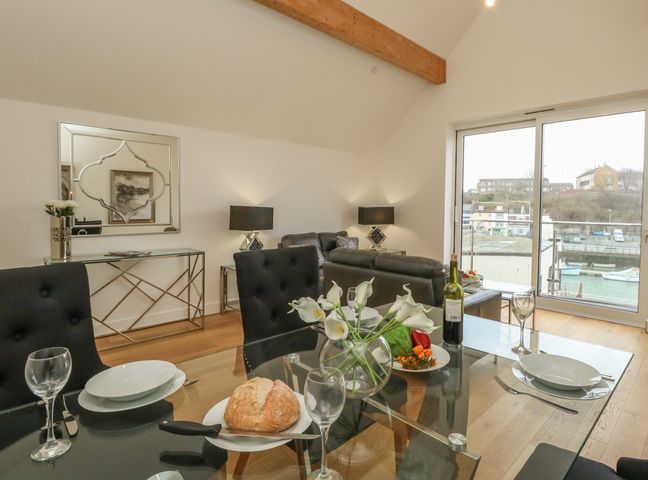 A dining area with a glass table set with plates wine glasses bread and flowers overlooking a living room with black sofas and large window at Harbourside Haven Penthouse 2 in Weymouth