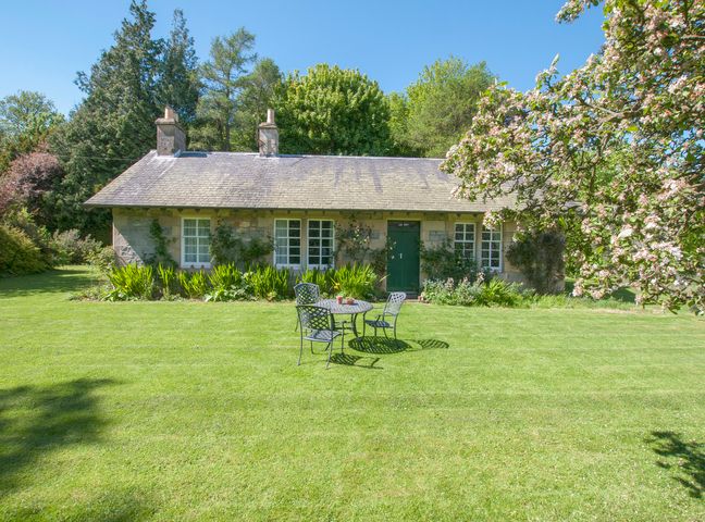 A stone cottage with green door and windows surrounded by grass and trees with outdoor table and chairs in front at Middle Cottage - Tarvit in Cupar Fife