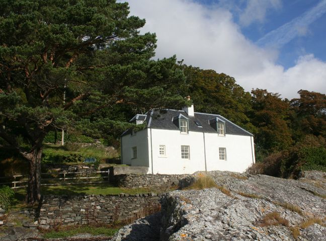 A white cottage surrounded by trees and stone walls with rocky terrain in the foreground at Craggan Cottage Balmacara Kyle Of Lochalsh