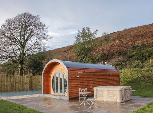 A wooden pod cabin with a round glass door a small patio table and chairs and a hot tub outside on a paved area at Stapeley Pod in White Grit near Chirbury